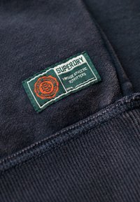 Sweat-shirt en tissu bleu marine avec une étiquette tissée verte et orange portant les inscriptions "Superdry" et "Vintage Athletic Outfitters", avec une texture côtelée.