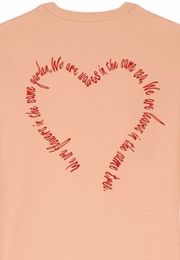 Persikų spalvos medvilninė marškinė su raudonais tekstais, išdėstytais širdies forma. Tekste yra citata apie vienybę ir ryšį. Apvalus iškirpimas.