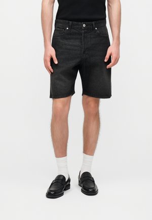 Mann trägt schwarze Jeans-Shorts, weiße Kniestrümpfe und schwarze Ledermokassins und steht vor einem schlichten Hintergrund.