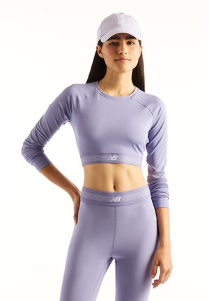 Femme portant un crop top à manches longues violet clair, un legging assorti et une casquette violet clair, posant avec une main sur la hanche sur fond blanc.
