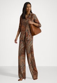 Long, patterned jumpsuit in warm tones of brown, orange, and blue with a collar and button front. Includes a brown handbag and heeled sandals.