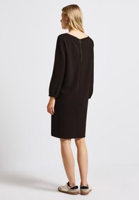 Femme aux cheveux blonds portant une robe noire à manches longues, longueur genoux, avec fermeture éclair au dos, debout et de dos, portant des baskets blanches.
