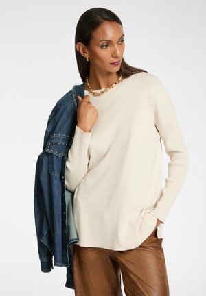 Ecrufarbener Pullover mit gerippter Textur, kombiniert mit einer Jeansjacke. Goldene Halskette als Akzent und lockere Passform. Braune Hose mit glänzender Oberfläche.
