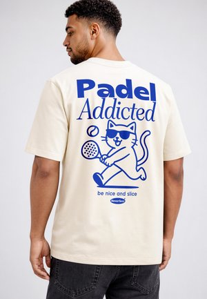 Man wearing cream T-shirt with blue graphic of cat playing padel and text "Padel Addicted, be nice and slice."
