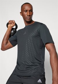 T-shirt de sport noir avec un motif texturé sur le devant, à manches courtes et à encolure ronde. Porté avec un pantalon de sport noir, arborant un logo.