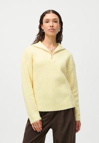 Yellow ribbed sweater with a quarter-zip collar and long sleeves. Worn over brown trousers. Textured fabric and relaxed fit.