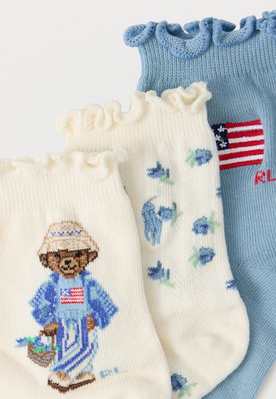 Τρεις κάλτσες με βολάν: μία μπλε με τη σημαία των ΗΠΑ, μία λευκή με μπλε λουλούδια και σκύλο, και μία λευκή με αρκούδα που φοράει καπέλο και κρατά καλάθι.