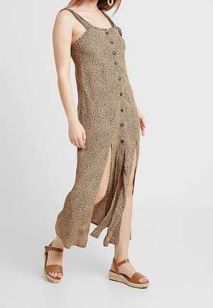 Vrouw die een mouwloze, bruin-zwart gevlekte midi-jurk met knopen, zijslitten en bruine sleehakken met enkelriempje draagt, loopt.