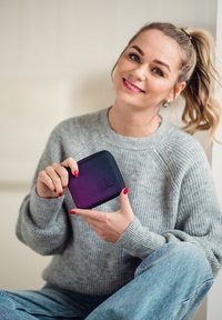 Compact wallet in gradient shades of blue and purple, smooth material, zip closure, embossed logo, held in a hand, with a knitted sweater backdrop.