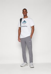 Tricou alb cu logo și grafică albastră Columbia, asociat cu pantaloni gri. Adidași gri texturați completează ținuta. Rucsac negru pe umăr.