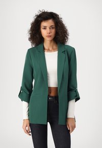 Grüner maßgeschneiderter Blazer mit lockerer Passform, leicht aufgerollten Ärmeln und zwei vorderen Taschen, getragen über einem weißen Cropped-Top und dunklen Jeans.