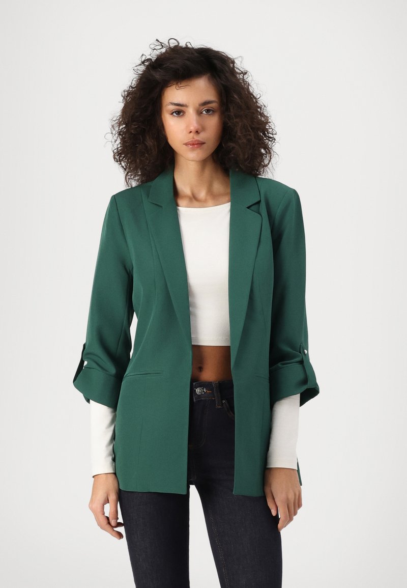 Grüner maßgeschneiderter Blazer mit lockerer Passform, leicht aufgerollten Ärmeln und zwei vorderen Taschen, getragen über einem weißen Cropped-Top und dunklen Jeans.