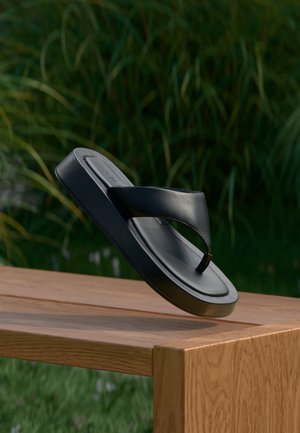 Schwarze Lederflip-Flop-Sandale, die über einer Holzbank im Freien schwebt, mit hohem grünem Gras unscharf im Hintergrund.