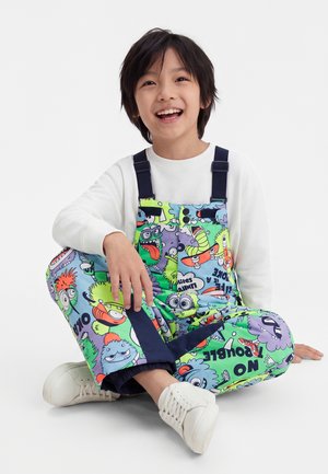 Lachend kind zittend met gekruiste benen, gekleed in een wit shirt met lange mouwen, kleurrijke tuinbroek met cartoonprint en witte sneakers op een witte achtergrond.