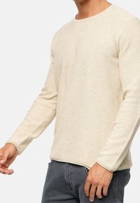 Un pull en tricot beige à manches longues avec un motif texturé, un col rond et des manches ajustées, associé à un pantalon gris.