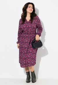 Vestido de manga larga, morado, con un patrón geométrico de corazones, cintura ceñida y a la altura de la rodilla. Combinado con un bolso negro y botas negras con tachuelas.
