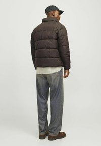 Chaqueta de plumas marrón con un alto cuello y diseño acolchado, llevada sobre una camisa de cuadros. Combinada con jeans azules holgados y zapatos marrones.