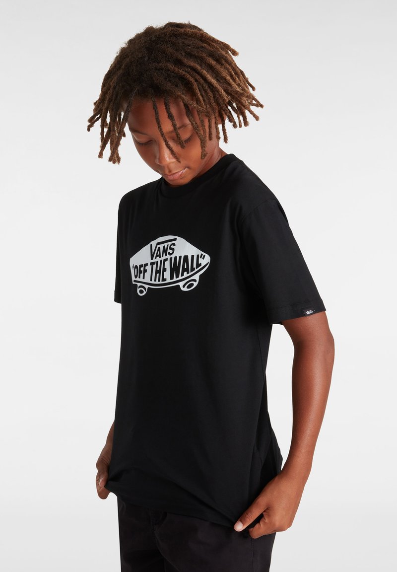 Camiseta negra de algodón, con un gráfico blanco de una patineta y el texto "VANS OFF THE WALL". Manga corta y escote redondo.