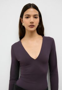 Dlhý rukáv body suit v tmavofialovej látke s výstrihom do V, priliehavým strihom a hladkou textúrou. Výrazná bezšvová konštrukcia a nenápadný štýl.
