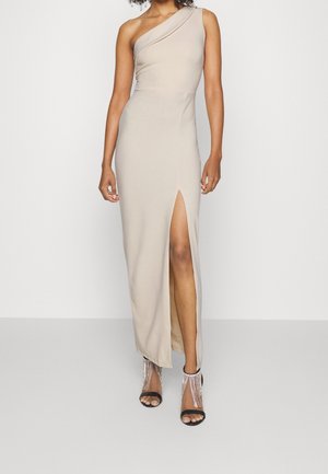 Femme portant une longue robe beige asymétrique avec une fente haute sur la cuisse et des sandales noires à talons hauts avec des brides ornées de strass.