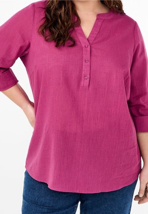 Woman wearing a long-sleeve pink buttoned blouse with rolled cuffs and blue jeans, hand on hip, against a white background.