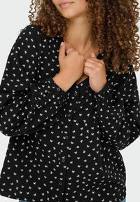 Zwarte blouse met knoopsluiting, voorzien van een subtiel wit strikpatroon, lange pofmouwen en een lichte textuur. Knopen langs de voorkant.