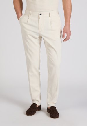 Mann trägt elfenbeinfarbene, schmal geschnittene Hose mit vorderen Falten und braune Wildleder-Loafer, steht mit einer Hand in der Tasche.