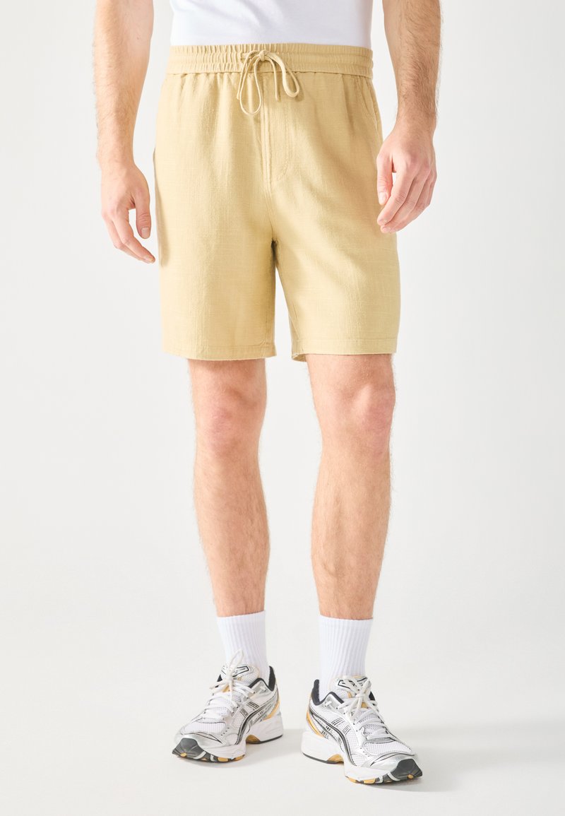 Beige bomulds shorts med elastisk talje og snøre. Har sidelommer, er midt-lår længde og har et tekstureret stof.
