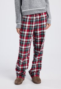 Persoon in een rode, groene en witte geruite pyjamabroek, een grijze trui met witte strepen en bruine slippers op een witte achtergrond.