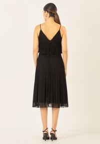 Schwarzes, gefaltetes Kleid mit V-Rücken, dünnen Trägern und einem mittellangen Rock. Mit einer dezenten transparenten Schicht am Saum. Einfaches und elegantes Design.