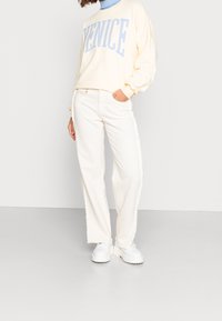 Felpa crema con testo blu "VENICE", abbinata a jeans bianchi a gamba larga. Calzature con suola spessa bianca. Tessuto morbido.
