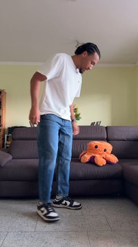 Une personne en t-shirt blanc et jeans bleus se tient à côté d'un canapé marron avec un jouet pieuvre en peluche orange et violet posé dessus.