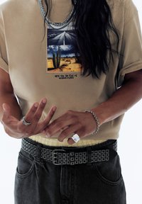 Beige t-shirt med et fotografisk tryk af en ørken scene, der inkluderer en kaktus. Båret med et mønstret bælte, mørke jeans og sølv tilbehør.