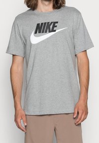 Muž v šedém tričku Nike s černo-bílým logem, ve světlých kalhotách, stojící na pozadí bez vzoru.