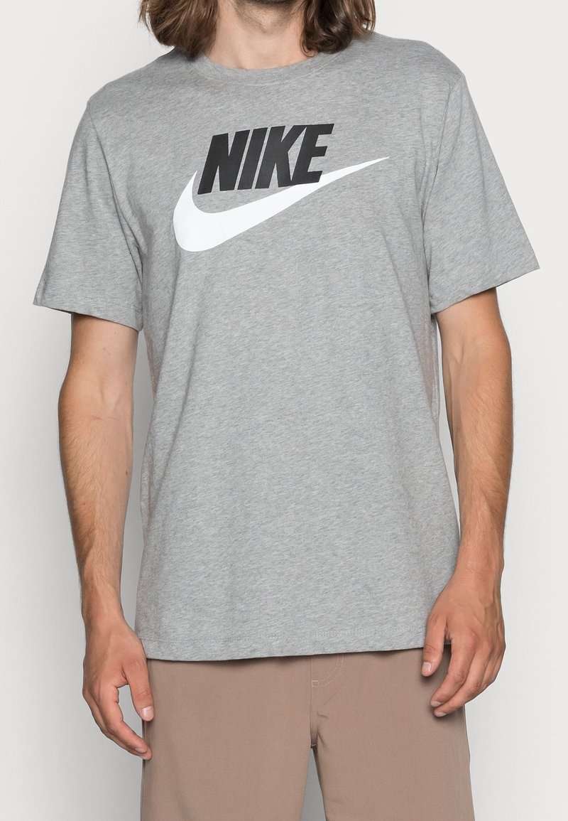 Muž v šedém tričku Nike s černo-bílým logem, ve světlých kalhotách, stojící na pozadí bez vzoru.