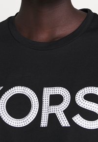 Musta t-paita, jossa on suuret, valkoisilla strasseilla koristellut kirjaimet "YORS". Kaulus on pyöreä ja istuvuus on normaali.