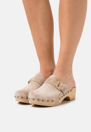 Beżowe zamszowe klapki na drewnianej platformie z ozdobną złotą klamrą. Akcenty w postaci ćwieków zdobią brzeg buta, projekt z otwartym tyłem.
