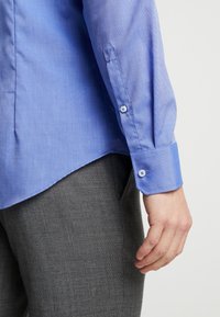 Světle modrá, texturovaná košile s manžetami na knoflíky. Obsahuje zúžený lem a hladkou látku. Šedé texturované kalhoty částečně viditelné.