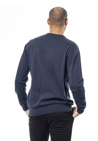 Homme tourné de dos portant un pull bleu marine à manches longues et un pantalon noir, les mains reposant sur les hanches, sur fond blanc.