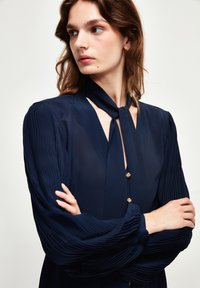Blusa navy con maniche plissettate, dettaglio del nodo al collo e bottoni dorati. Il tessuto trasparente offre una morbida texture e un'elegante silhouette.