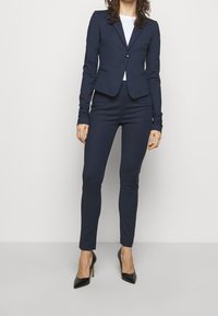 Marineblauwe tailored blazer met een enkele knoop, vergezeld van slim-fit marineblauwe broek. Glad materiaal met lange mouwen en spitse zwarte hakken.