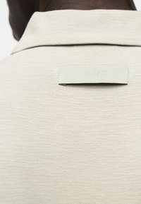 Κοντινό πλάνο στο άνω μέρος της πλάτης ενός ατόμου που φορά ένα ανοιχτό μπεζ υφασμάτινο πουκάμισο με μια μικρή ορθογώνια ετικέτα με ανάγλυφο το «HUGO» κάτω από το γιακά.