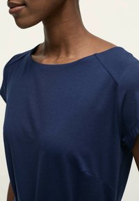 Top bleu marine à manches courtes avec un col rond, texture lisse et un détail de poche latérale subtil. Présente une coupe décontractée.