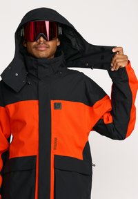 Veste de ski noire et orange avec un col haut et une capuche amovible, dotée de deux poches avant et d'un tissu texturé. Lunettes de ski teintées en rouge.