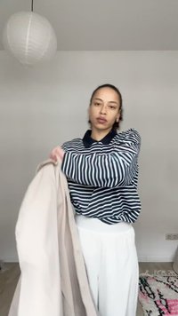 Žena oblečená v čierno-bielom pruhovanom tričku a bielych nohaviciach, držiaca béžovú bundu v minimálne zariadenej miestnosti s visiaci lampou.