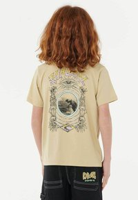 Camiseta beige con un diseño gráfico en la parte posterior, que incluye un motivo de ojo, una imagen central y elementos decorativos en negro, amarillo y púrpura.