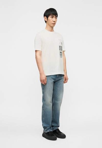 Νεαρός άνδρας όρθιος, φοράει λευκό μπλουζάκι με σχέδι ο, ανοιχτό μπλε τζιν και μαύρα αθλητικά, μπροστά σε απλό λευκό φόντο.