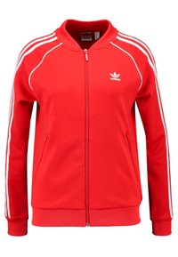 Červená bunda na zip z měkké látky, s bílým designem tří pruhů podél rukávů a malým bílým logem na hrudi.