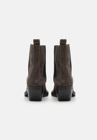 Botas de tobillo de ante marrón con paneles laterales elásticos y un tacón negro robusto. Presentan una textura suave y tiradores en el talón para facilitar su uso.