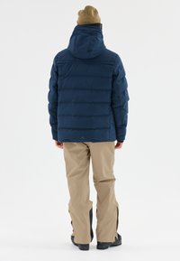Veste en duvet marine avec capuche, comportant des sections matelassées. Assortie à un pantalon beige imperméable et des chaussures noires, vue de dos.